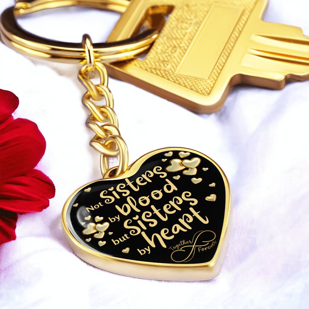 Gold-toned heart-shaped keychain with a black background and printed message: 'Not Sisters by blood, but Sisters by heart.' Small gold hearts surround the message. in white background , next to a red flower