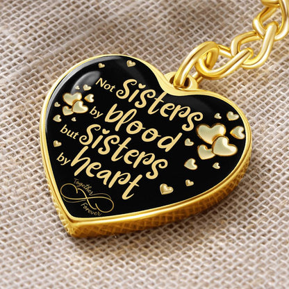 Close look to a Gold-toned heart-shaped keychain with a black background and printed message: 'Not Sisters by blood, but Sisters by heart.' Small gold hearts surround the message