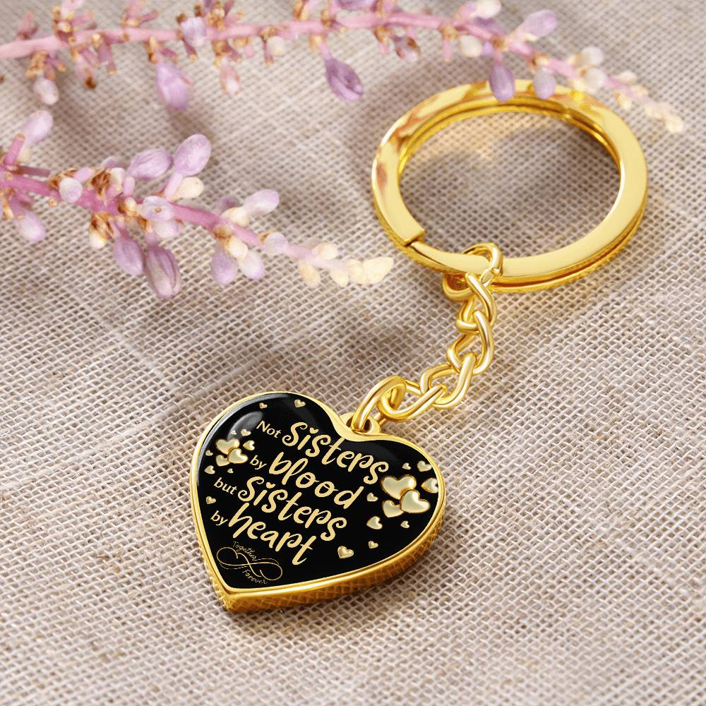 Gold-toned heart-shaped keychain with a black background and printed message: 'Not Sisters by blood, but Sisters by heart.' Small gold hearts surround the message. in floral background