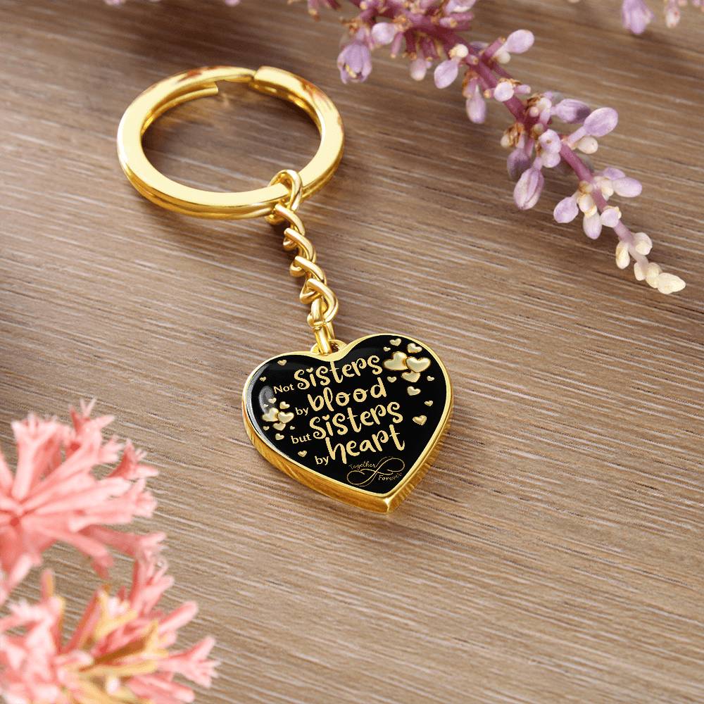 Gold-toned heart-shaped keychain with a black background and printed message: 'Not Sisters by blood, but Sisters by heart.' Small gold hearts surround the message, in a brown background