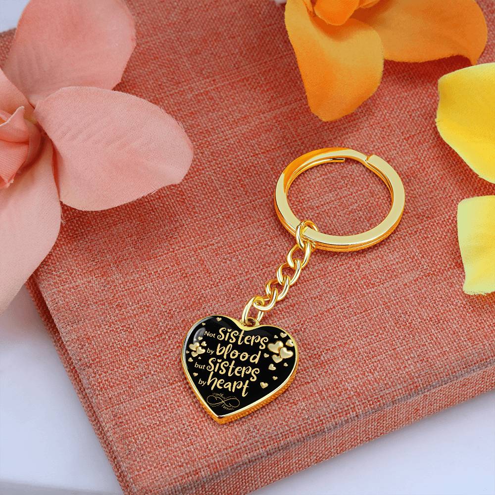 Gold-toned heart-shaped keychain with a black background and printed message: 'Not Sisters by blood, but Sisters by heart.' Small gold hearts surround the message. set on a orange napkin