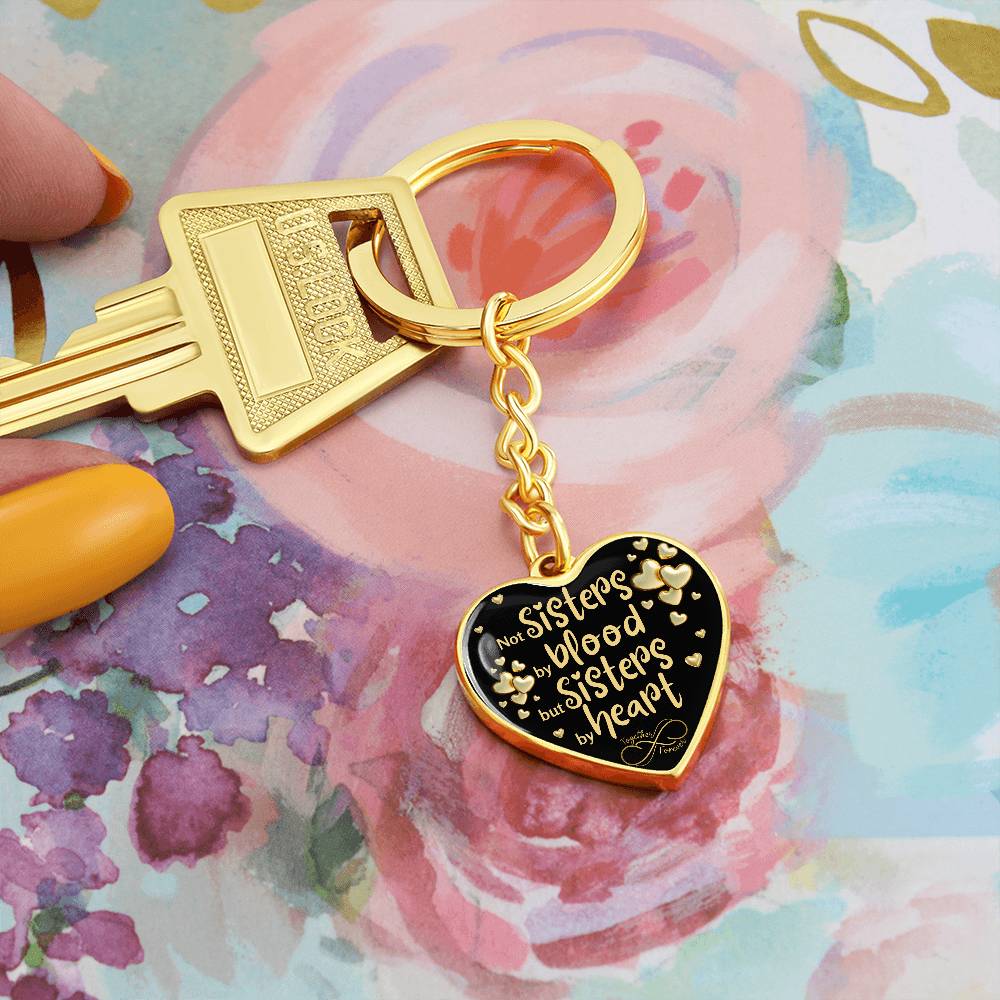 Gold-toned heart-shaped keychain with a black background and printed message: 'Not Sisters by blood, but Sisters by heart.' Small gold hearts surround the message. in a beautifull floral background.