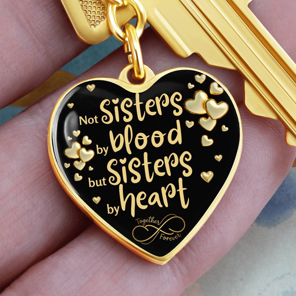 Human fingers holding a Gold-toned heart-shaped keychain with a black background and printed message: 'Not Sisters by blood, but Sisters by heart.' Small gold hearts surround the message