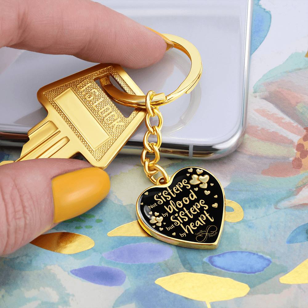 Woman hand holding a Gold-toned heart-shaped keychain with a black background and printed message: 'Not Sisters by blood, but Sisters by heart.' Small gold hearts surround the message