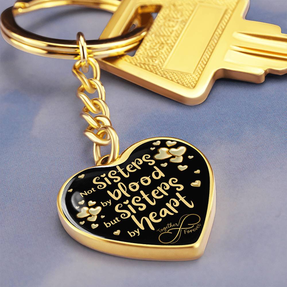 Gold-toned heart-shaped keychain with a black background and printed message: 'Not Sisters by blood, but Sisters by heart.' Small gold hearts surround the message. in blue background