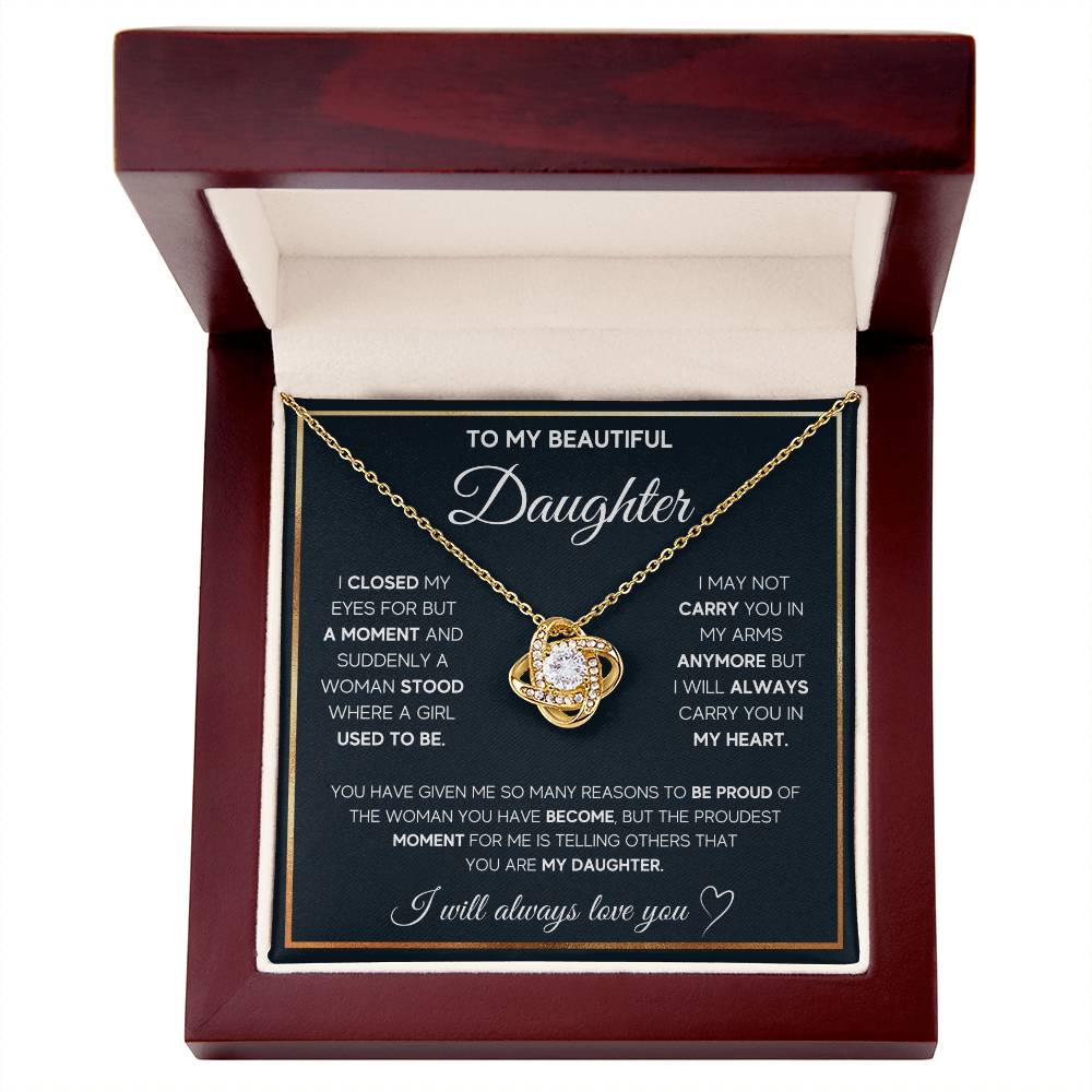 A gold necklace with a knot-shaped pendant in a wooden box, a gift from a parent to their daughter with a heartfelt message.I n white background