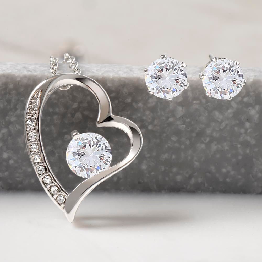 A close-up image of a heart-shaped necklace and matching earrings. The necklace has a larger center cubic zirconia stone and smaller stones along the heart's outline. The earrings are stud earrings with cubic zirconia stones. 